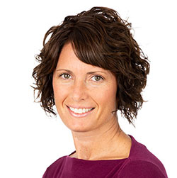 A woman with short, curly brown hair wears a burgundy top. Her face is intentionally blurred, and the plain white background suggests a professional or clinic-related setting, such as a hearing clinic.