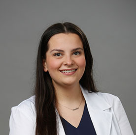 A professional wearing a white lab coat over a navy blue top stands against a plain gray background, suggesting a healthcare setting such as a hearing clinic. No text present.