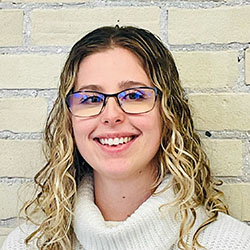 A woman with curly, shoulder-length blonde hair wears a white knit turtleneck sweater. She stands against a beige brick wall backdrop. Her face is intentionally blurred for privacy. No visible text or branding.