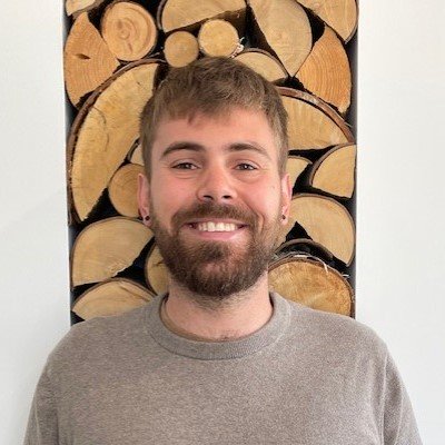 A person in a gray sweater stands in front of a neatly stacked wooden log display arranged in a decorative pattern against a white background. The face is blurred.