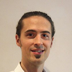 Andy Berezowsky, hearing care professional with long tied-back hair, wearing a white shirt against a neutral background.