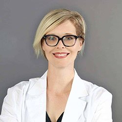 A blonde woman wearing a white lab coat is standing confidently against a plain gray background, likely representing a professional audiologist or hearing specialist at a hearing clinic. No text visible.