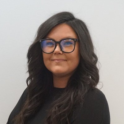 A person with long, wavy black hair wearing a black top stands against a plain, light-colored background. Their face is blurred, offering anonymity. No text or hearing aids are visible.