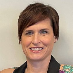 A professional with short brown hair wears a black-collared outfit featuring floral patterns on the shoulder. The setting is a neutral, beige indoor background, consistent with a hearing clinic environment. No visible hearing aids or identifiable branding.