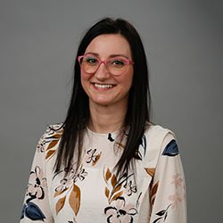 A person with shoulder-length dark hair wears a floral-patterned blouse and a discreet pink hearing aid, standing against a plain gray backdrop, likely in a hearing care or clinic-related context.