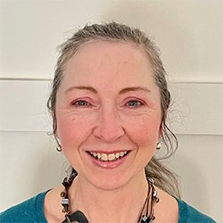 A person with grayish-brown hair tied back is wearing a teal top and a beaded necklace. The background features a plain, off-white wall. The face is intentionally blurred. No text or hearing aid branding is visible.