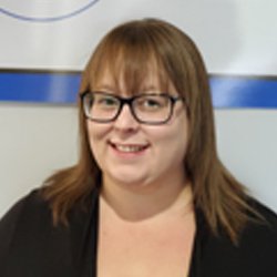 A person with shoulder-length brown hair, wearing a black top, stands indoors against a white and blue banner. The image is related to HearingLife, a hearing care clinic.