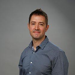 A person wearing a blue patterned button-up shirt stands against a plain gray background, with their face blurred for anonymity. There is no visible text or branding present.
