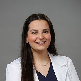 A professional wearing a white lab coat over a navy blue top stands against a plain gray background, suggesting a healthcare setting such as a hearing clinic. No text present.