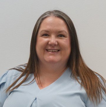 A person with long, straight brown hair is wearing a light blue blouse with a pleated neckline, set against a plain, light gray background. No text or identifiable hearing aids are visible.