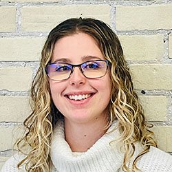A woman with curly, shoulder-length blonde hair wears a white knit turtleneck sweater. She stands against a beige brick wall backdrop. Her face is intentionally blurred for privacy. No visible text or branding.