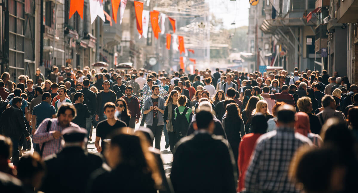 A dense crowd of people walks along a bustling urban street adorned with red flags. Buildings line each side, creating a lively setting under bright daylight. No text visible.