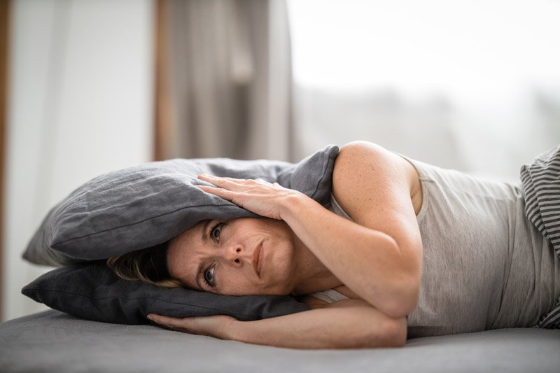 A person lies on their side in bed, holding a gray pillow over their head. The room features soft, natural lighting with neutral-toned bedding and curtains in the background.