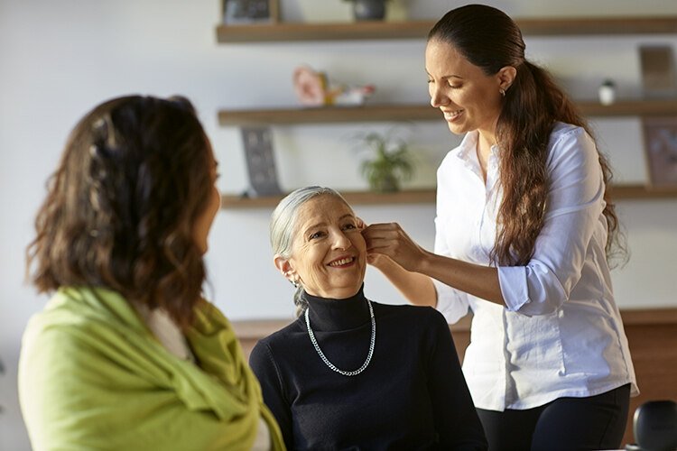 Hearing care specialist fitting a hearing aid on an older woman while another woman observes in a modern clinic setting.