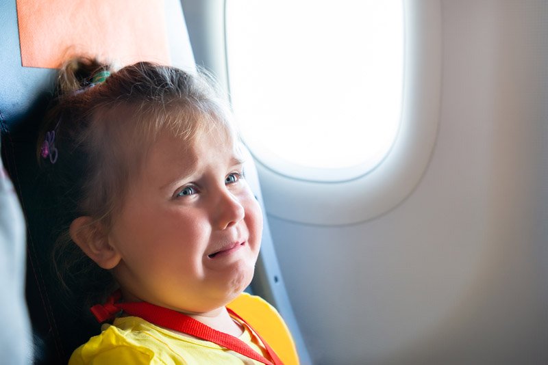 A young child, seated on an airplane, is wearing a yellow garment and a red lanyard. The child is near a window, with bright daylight visible outside.