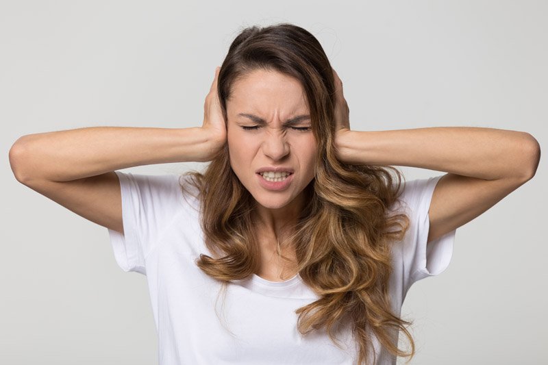 A woman, dressed in a white shirt, covers both ears with her hands, suggesting hearing strain or discomfort, set against a neutral gray background. No text or hearing device branding is visible.