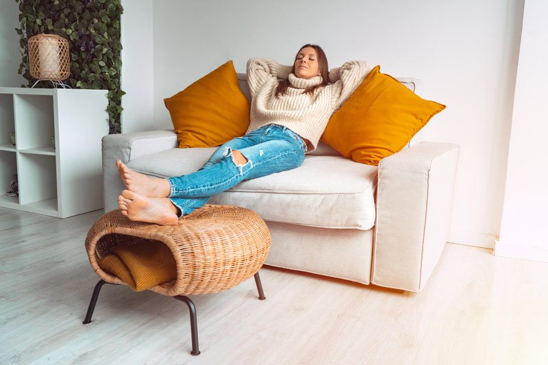 A person lounges barefoot on a beige sofa with orange throw pillows, wearing a knitted sweater and ripped jeans; a wicker ottoman with a folded blanket sits in a bright, minimalist room.