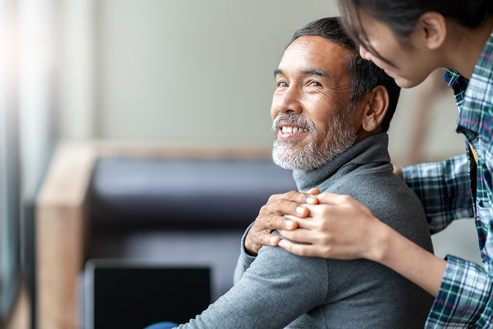 A seated individual wearing a gray turtleneck is gently comforted by someone placing supportive hands on their shoulders. The setting appears to be indoors with soft natural light near a couch.