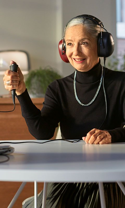 An individual undergoing a hearing test wears large over-ear headphones while holding a response button in a softly lit hearing clinic with modern decor and plants in the background.