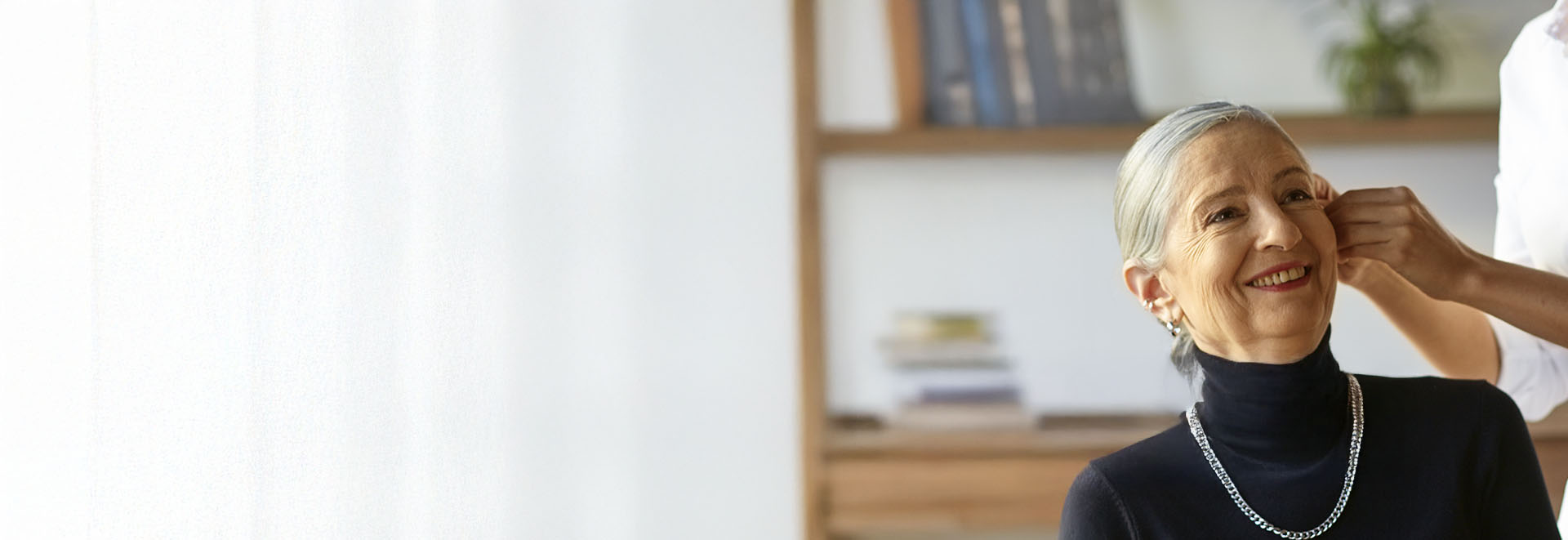 A senior individual wearing a black turtleneck and necklace sits indoors, while an unseen professional adjusts or fits a hearing aid. The background features bookshelves and soft lighting.