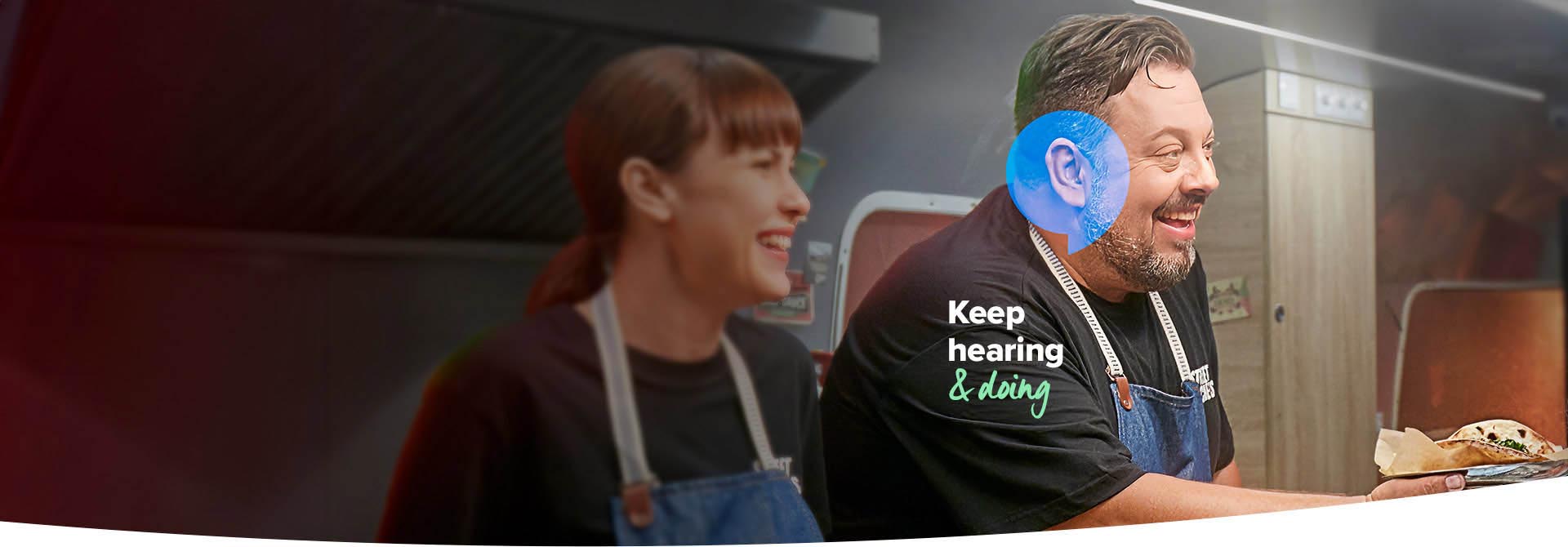 A person wearing a blue hearing aid serves food in a professional kitchen alongside a colleague. The text reads, ’Keep hearing & doing,’ emphasizing hearing care solutions in an active environment.