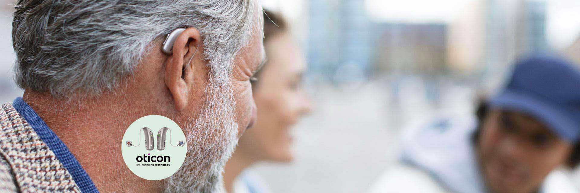 A senior man with gray hair wears an Oticon hearing aid behind his ear. In an outdoor urban setting, blurred pedestrian faces and buildings create a busy background. Text: ’Oticon, life-changing technology.’