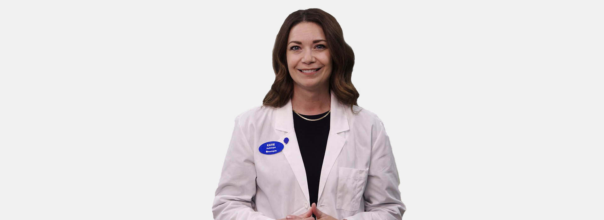 A female audiologist in a white lab coat stands against a plain white background. A blue name tag reads: 'Katie, Audiologist @HearingLife.' She appears professional and approachable, representing hearing care services.