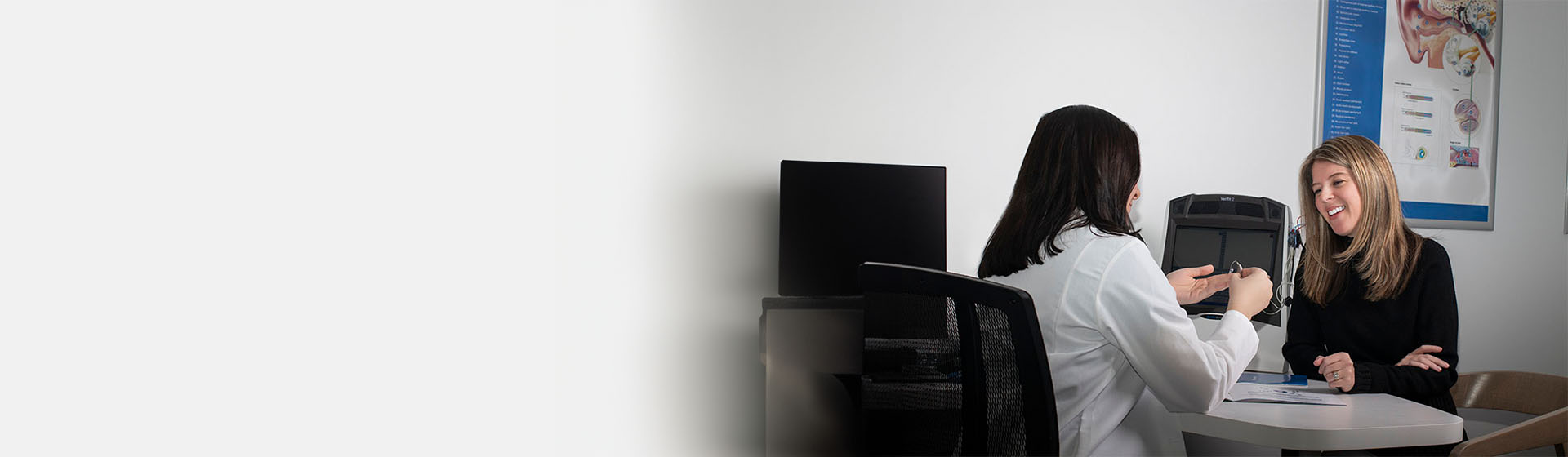 Audiologist shows a hearing aid to a client seated at desk in a clinic with educational ear anatomy posters on the wall.