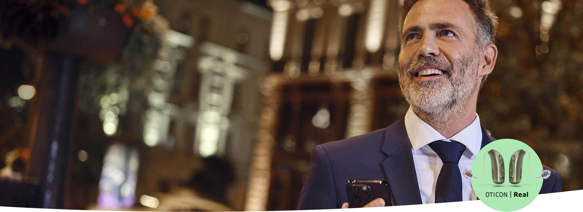 A man in a suit holds a smartphone in a dimly lit urban setting featuring blurred lights. The bottom-right corner displays ’Oticon | Real’ with images of Oticon Real hearing aids.