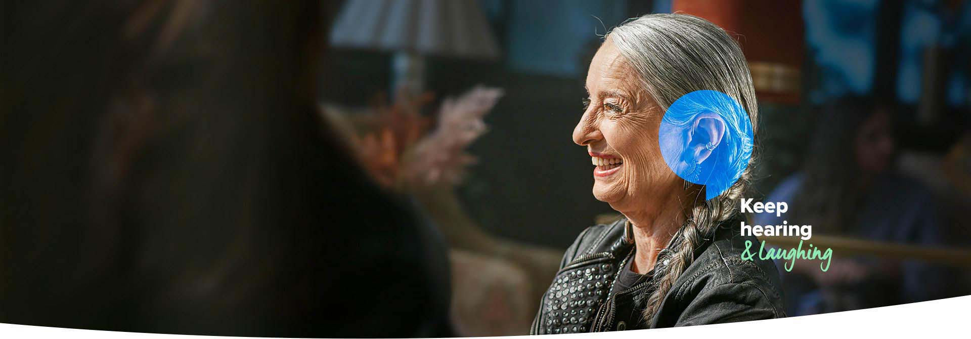 A senior individual with gray hair wears a black jacket with studded details, sitting in a warmly lit environment. Blue ear anatomy graphics highlight the ear. Text reads: ’Keep hearing & laughing.’
