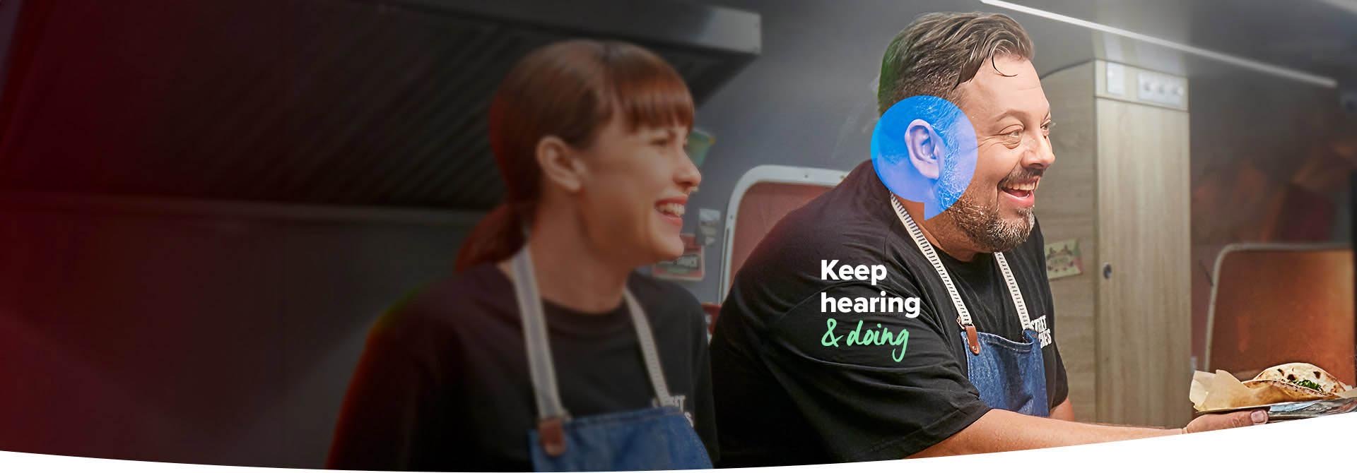 A person wearing a blue hearing aid serves food in a professional kitchen alongside a colleague. The text reads, ’Keep hearing & doing,’ emphasizing hearing care solutions in an active environment.