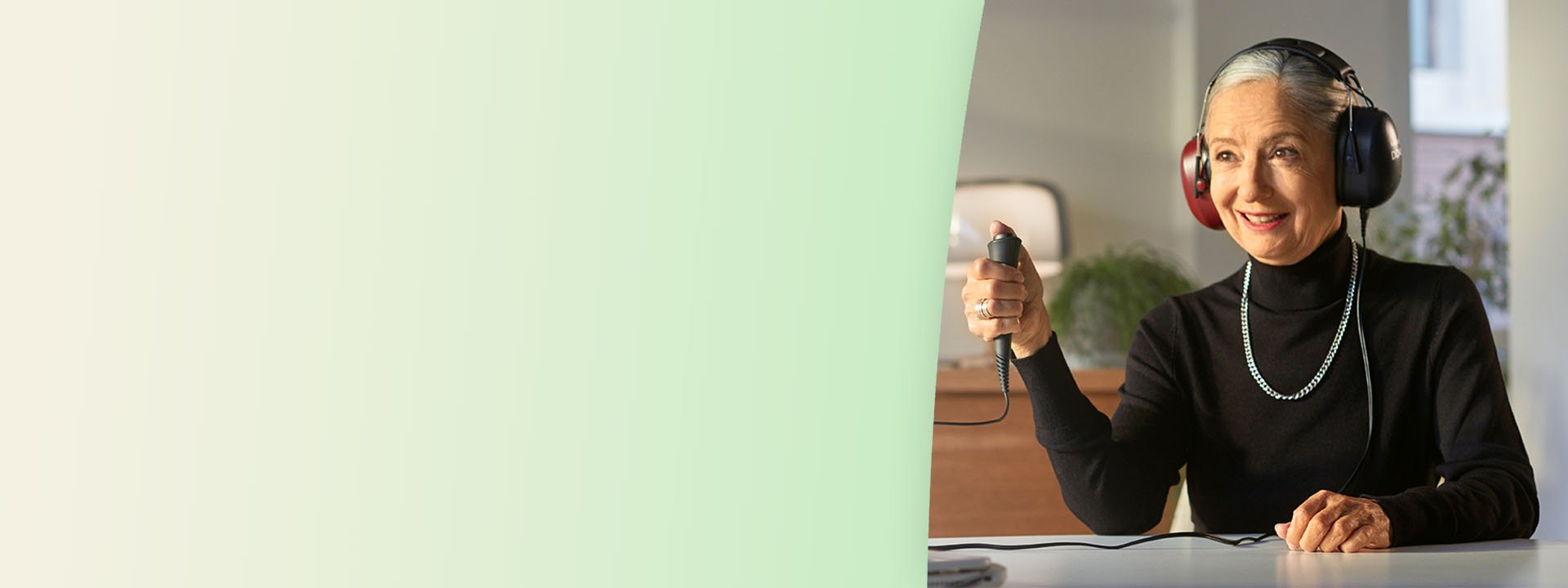 An individual undergoing a hearing test wears large over-ear headphones while holding a response button in a softly lit hearing clinic with modern decor and plants in the background.