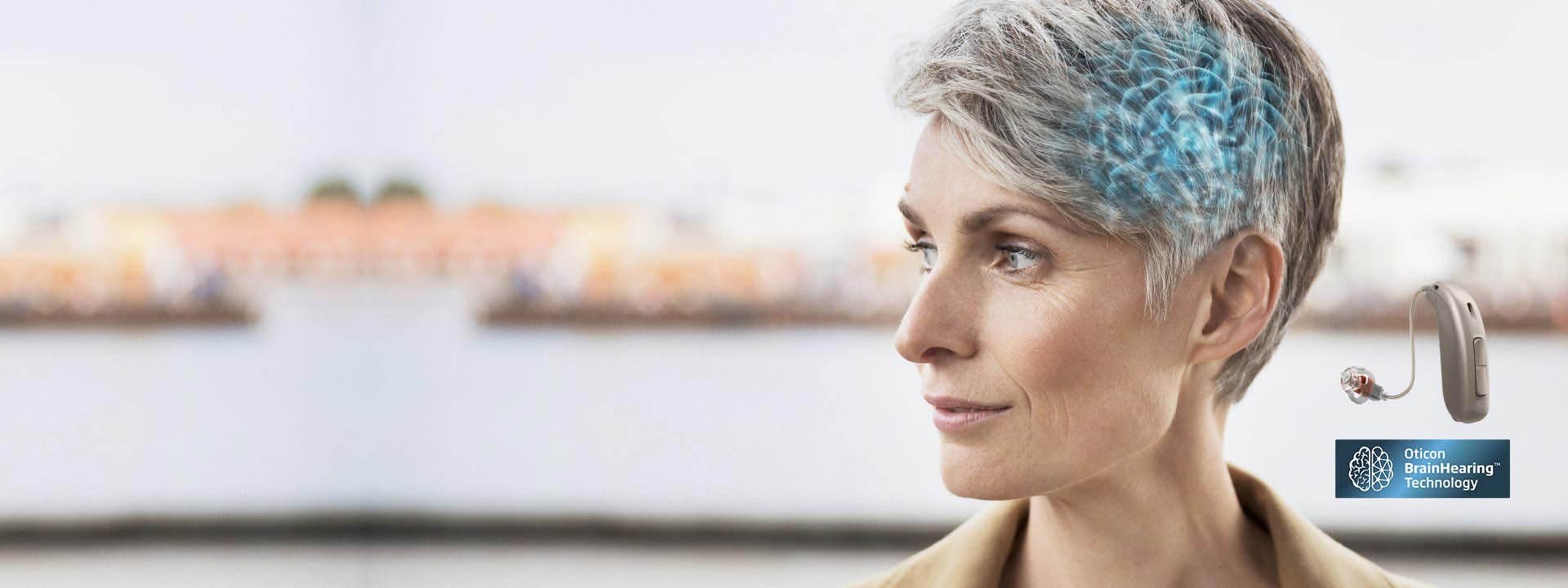 A gray-haired individual wears an Oticon hearing aid, with an overlay illustrating ’brainhearing technology.’ The background is blurred, showing waterfront buildings. Text: ’Oticon BrainHearing Technology’ with a brain icon.