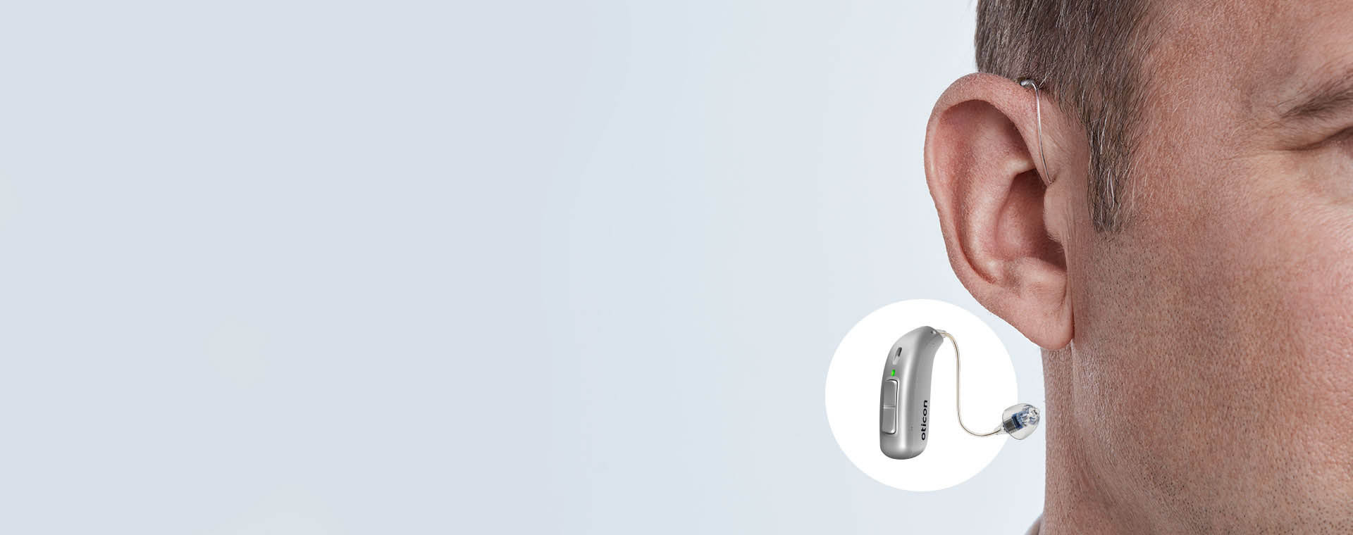 A man wears an Oticon hearing aid discreetly positioned behind his ear, with a close-up inset highlighting its sleek silver design and features, set against a light gray background.