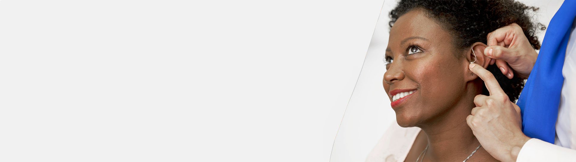 A clinician fits a hearing aid onto a person’s ear, adjusting it with care. The setting appears to be a professional hearing clinic, emphasizing personalized hearing care services. No visible text.