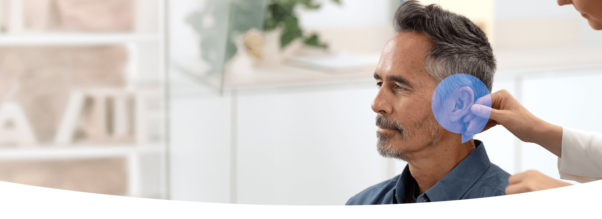 A hearing care professional carefully adjusts a hearing aid behind a man’s ear in a modern clinic with white furnishings, shelves, and greenery, reflecting a professional hearing test environment.