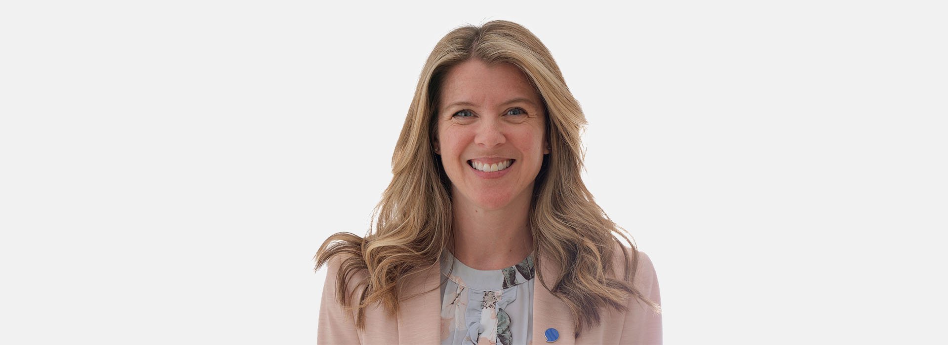 A woman with wavy blonde hair wears a light pink blazer and a patterned blouse, standing against a plain white background. Her face is blurred, and a blue circular pin is visible on her blazer lapel.