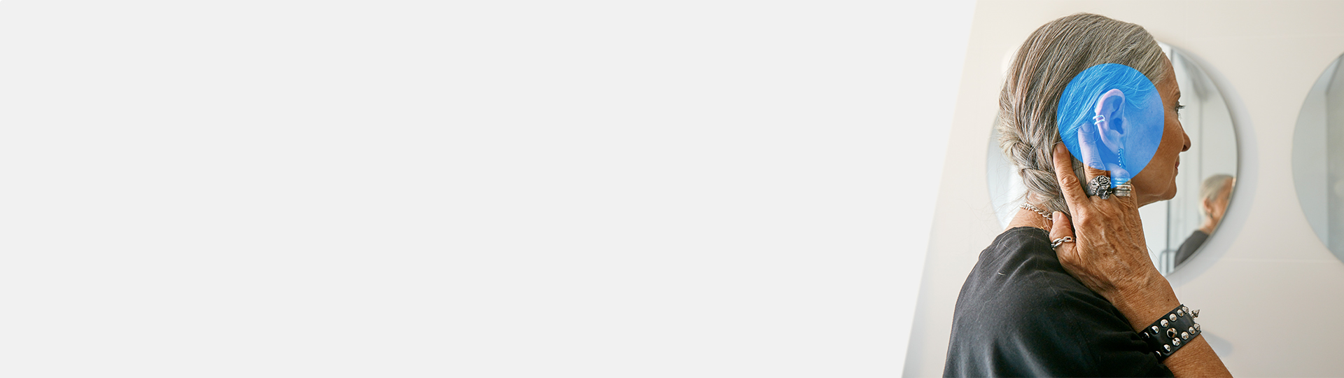 Senior woman with a hearing aid adjusting it near her ear while standing in front of a mirror.