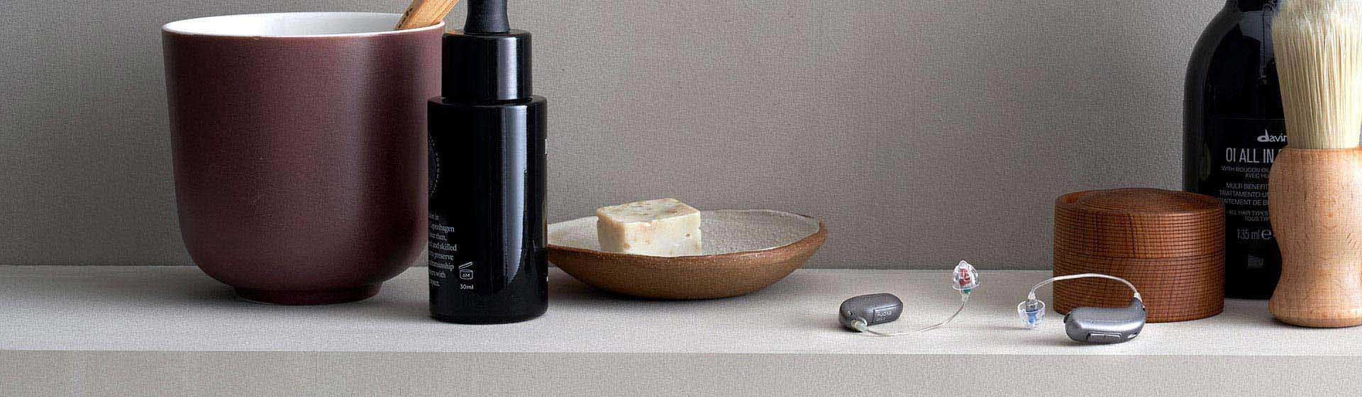 Two sleek hearing aids with clear earpieces rest on a neutral surface, surrounded by grooming items—a maroon mug, black bottle, wooden bowl, bar soap, brush, and wooden container. No text appears.