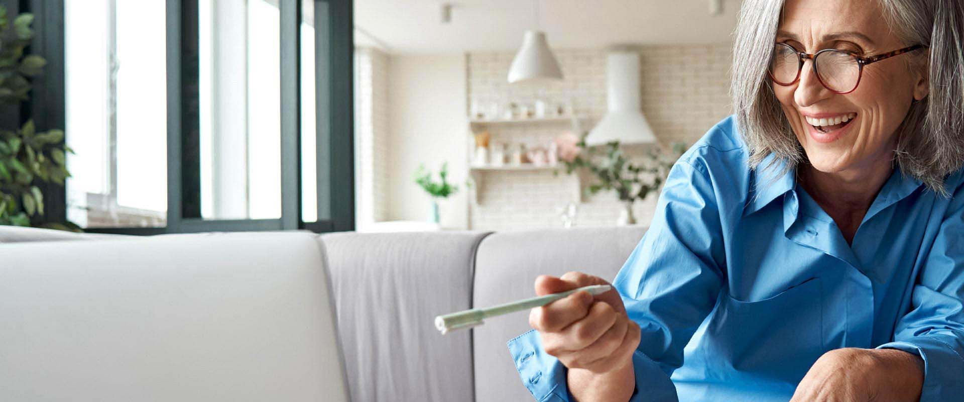 A person with shoulder-length gray hair, wearing a blue shirt, holds a small, slender object in a modern, well-lit living space with plants and shelves in the background.