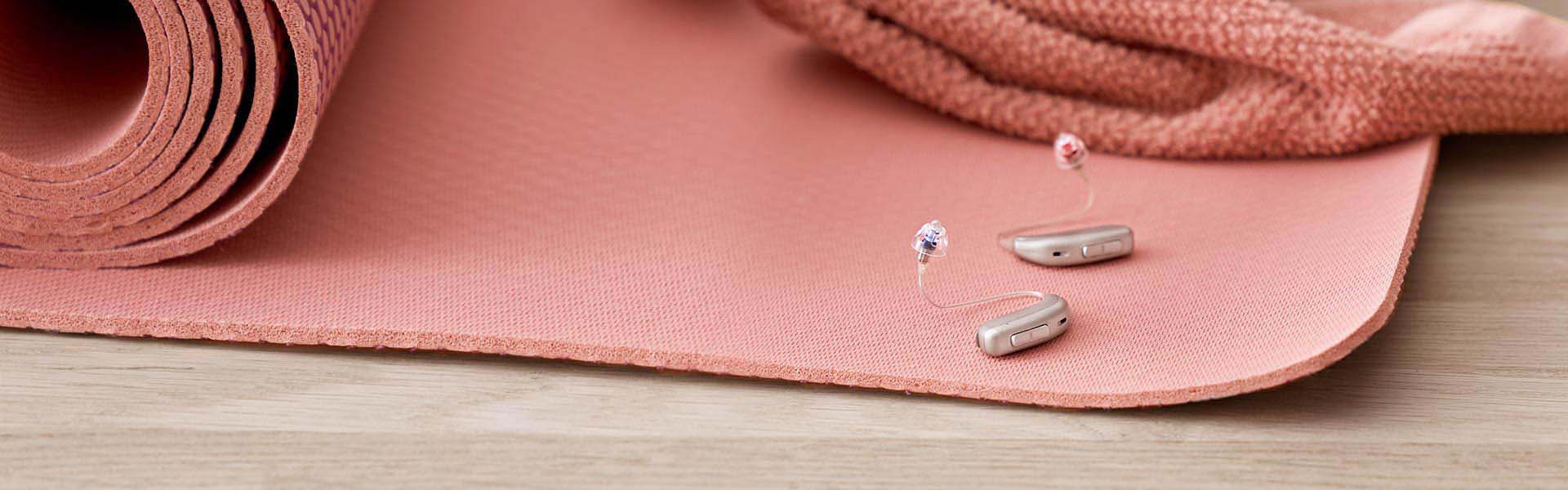 Two sleek beige hearing aids rest on a pink yoga mat, accompanied by a matching rolled towel, highlighting an active, health-focused lifestyle in a serene indoor setting.
