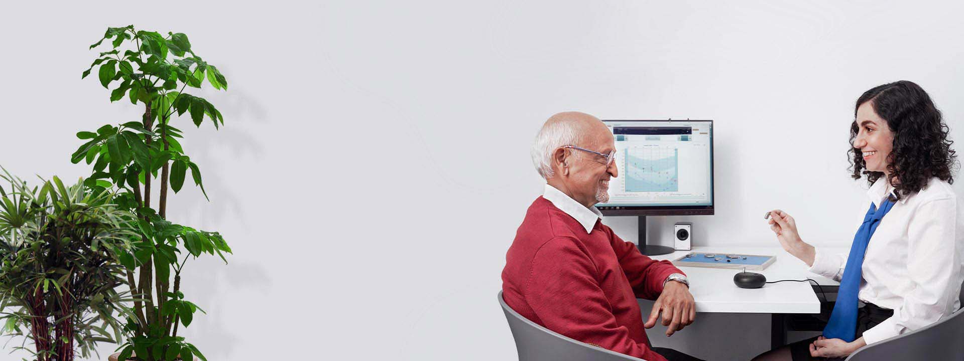 An older man in a red sweater sits across from an audiologist wearing a white shirt with a blue tie, reviewing a hearing test displayed on a computer monitor in a well-lit hearing clinic. Indoor plants are visible in the background, adding a calming touch.