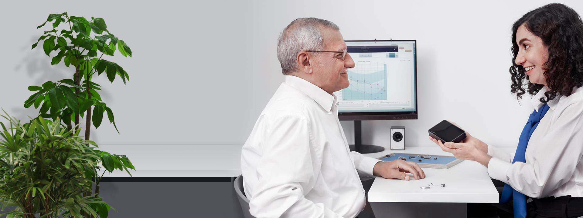 An older man sits with an audiologist in a hearing clinic. The audiologist holds a device, discussing hearing aids on the desk. A monitor displays hearing test results; plants decorate the scene.