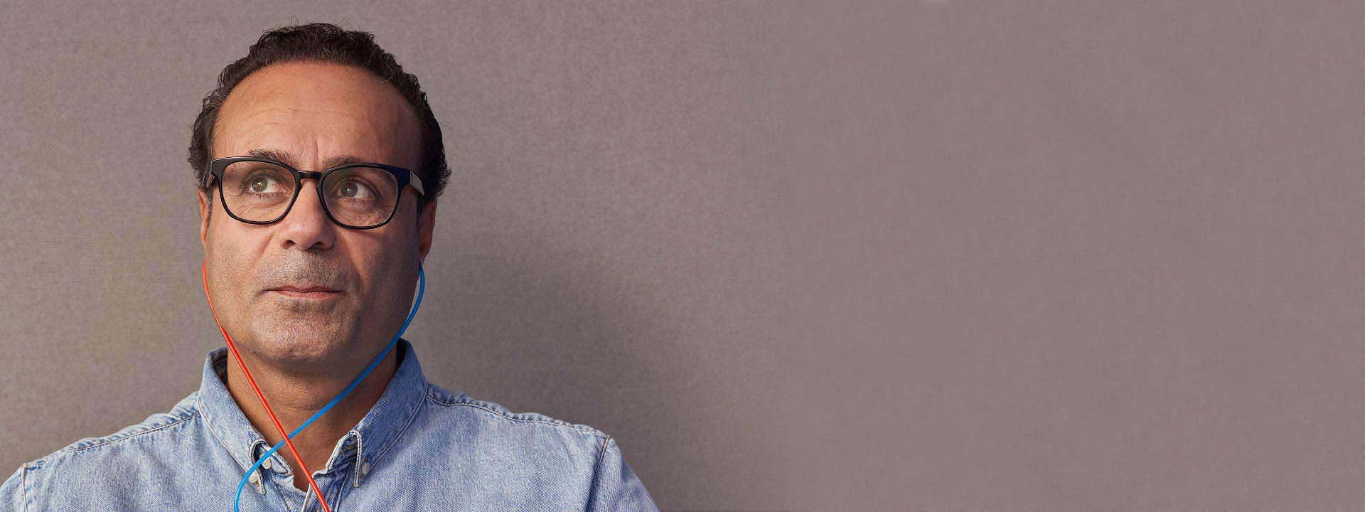 A person wearing a light denim shirt uses blue and red-tipped earphones for a hearing test against a plain, muted beige background. The context suggests a professional hearing care setting. No text is visible.