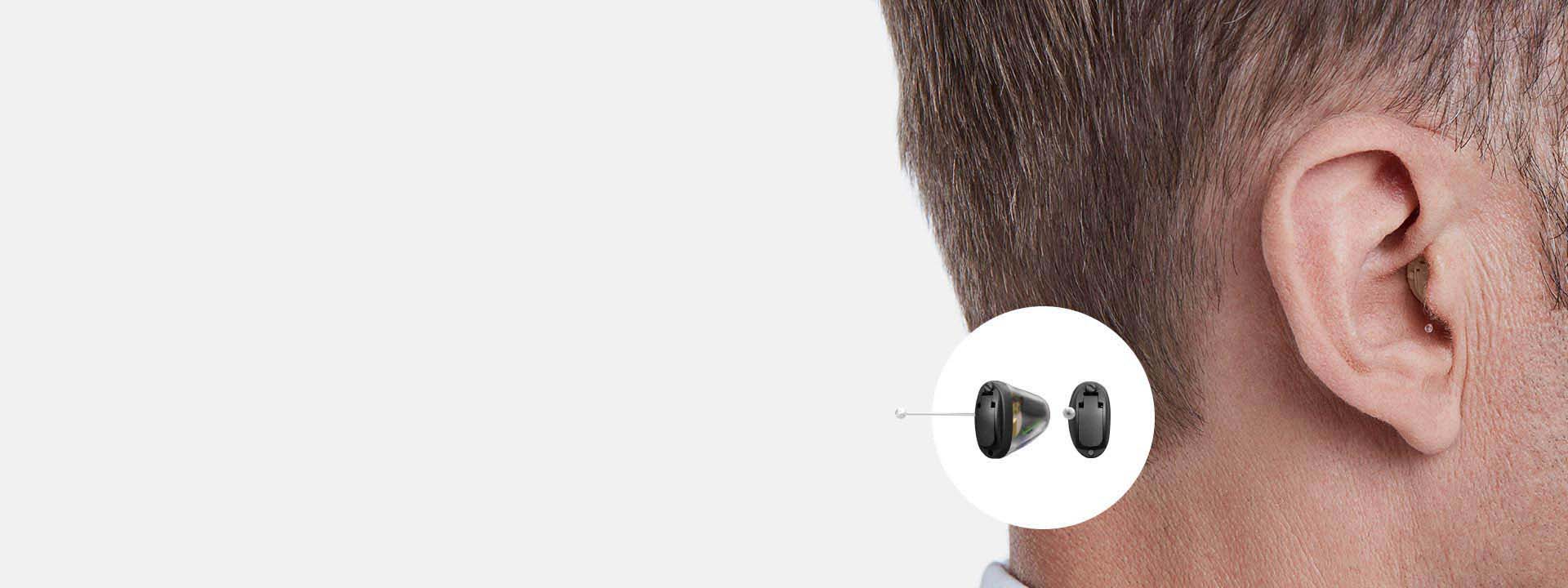 A discreet in-ear hearing aid is positioned in the ear canal of a man’s right ear, with a magnified inset showcasing the device’s design against a neutral background. No text is present.