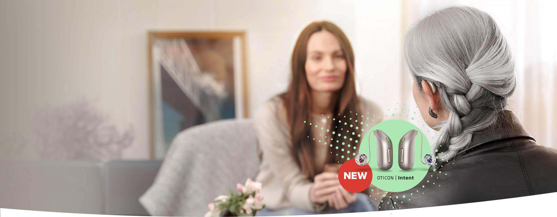 A woman with gray braided hair wears an Oticon hearing aid during a conversation in a cozy room. A highlighted banner promotes ’NEW Oticon | Intent’ hearing aids in the foreground.