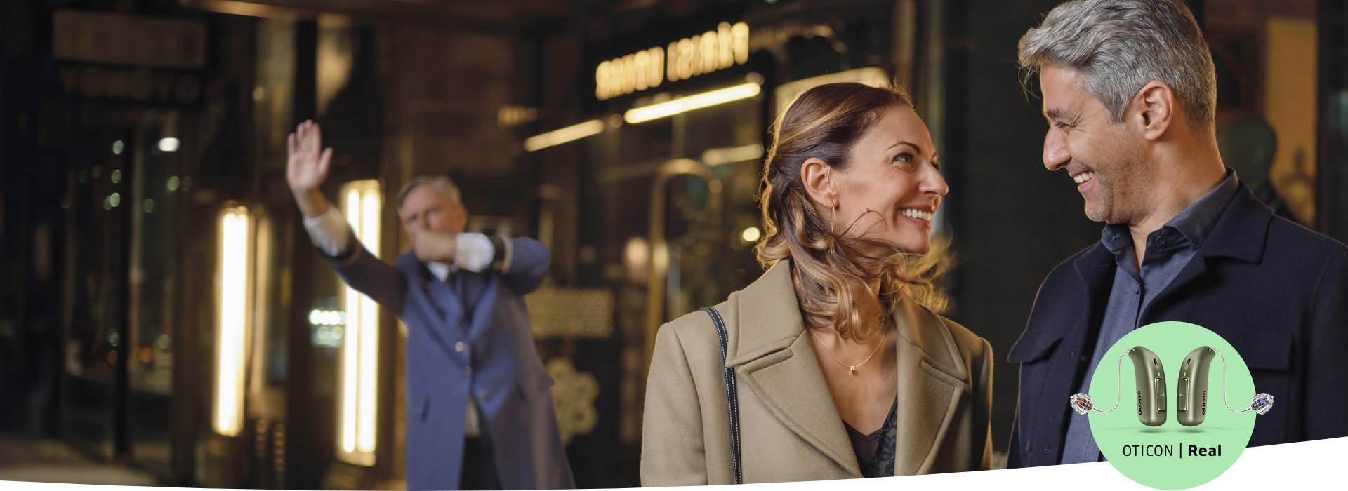 A couple stands outdoors near an illuminated urban storefront, engaging in conversation, while a doorman gestures in the background. The Oticon Real hearing aids logo is displayed prominently in the corner.