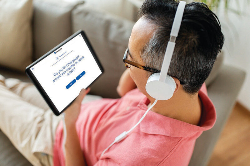 A man wearing white headphones views a tablet displaying a hearing test question from HearingLife. The text on the screen reads: ’Do you find that people around you mumble or speak quietly?’ He is seated on a beige couch in a well-lit room.