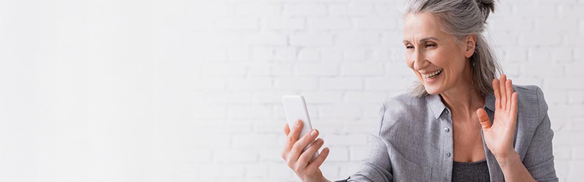 A gray-haired individual holds a smartphone while gesturing with their other hand, seated against a white brick background. The scene suggests digital communication or online consultation in a neutral setting.