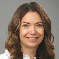 A person with long, wavy brown hair wears a white collared shirt. The background is a neutral gray, suggesting a professional setting, such as a hearing clinic. The face is intentionally blurred.